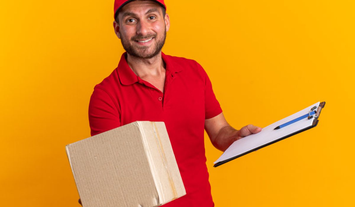 smiling-young-caucasian-delivery-man-red-uniform-cap-holding-clipboard-stretching-out-cardboard-box-towards-camera
