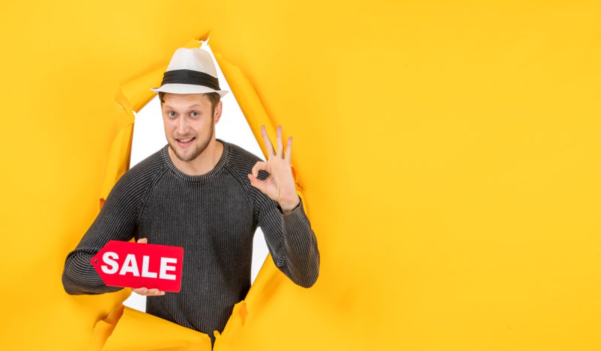 top-view-smiling-young-guy-holding-sale-sign-making-eyeglasses-gesture-torn-yellow-wall